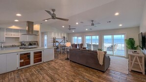 A spacious living area with a large window overlooking the beach, a cozy seating area, and a modern kitchen with white cabinets and wooden flooring. - Spacious Gulf Front Home on Extra Wide Lot! (Port St. Joe)