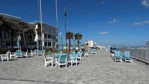 A beachfront promenade with rows of white lounge chairs and blue umbrellas, a palm tree-lined walkway, and a building with a green roof in the background. - On the beach (Daytona Beach)
