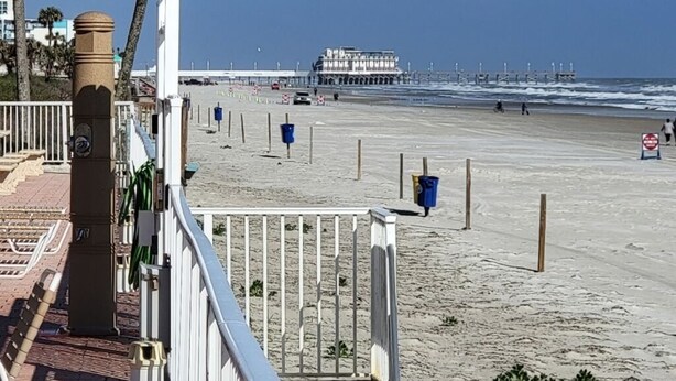A beachside walkway with a view of the ocean, a pier, and a building in the distance. - On the beach (Daytona Beach)