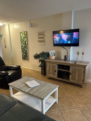 A living room with a television, a leather sofa, a coffee table, a cabinet, and a wall with a plant and a sign. - On the beach (Daytona Beach)