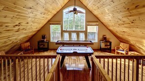 Acogedor cuarto ático con mesa de billar, paredes de madera y un ventanal con vistas a los árboles.