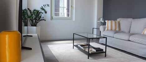 A modern living room with a glass coffee table, a sofa with striped pillows, a potted plant, and a window with shutters.