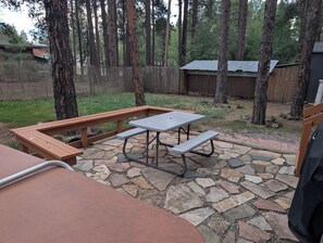 En stenbelagt terrasse med et picnicbord og bænke, omgivet af træer og et træhegn.