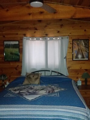 Une pièce de style cabane en bois, avec un lit, un ventilateur de plafond et deux tableaux encadrés accrochés aux murs.