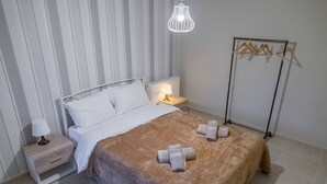 A neatly made bed with a brown blanket and two towels, a bedside table with a lamp, a radiator, and a clothing rack. - Historic Luxury House Heart of Meteora (Kalabaka)