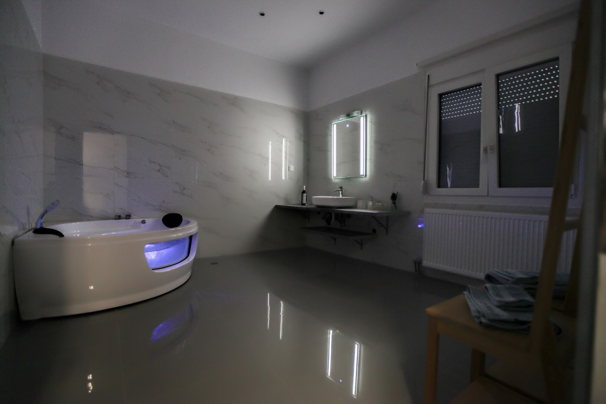 Ein modernes Badezimmer mit Marmorwand, einer eingebauten Badewanne mit Lichtfunktion, einem Waschtisch mit Spiegel und einem Fenster mit Jalousien.