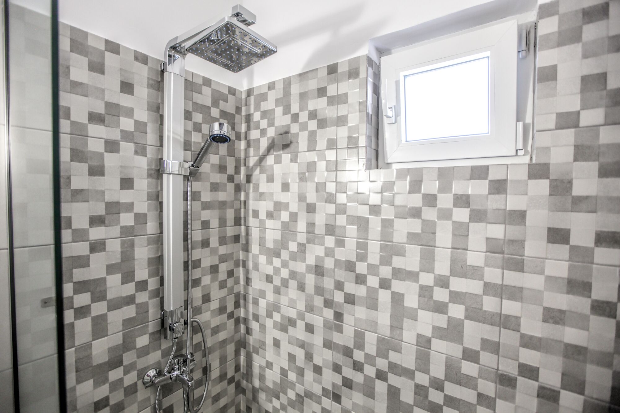 A shower area with a checkered tile wall, a handheld showerhead, and a window above the shower.