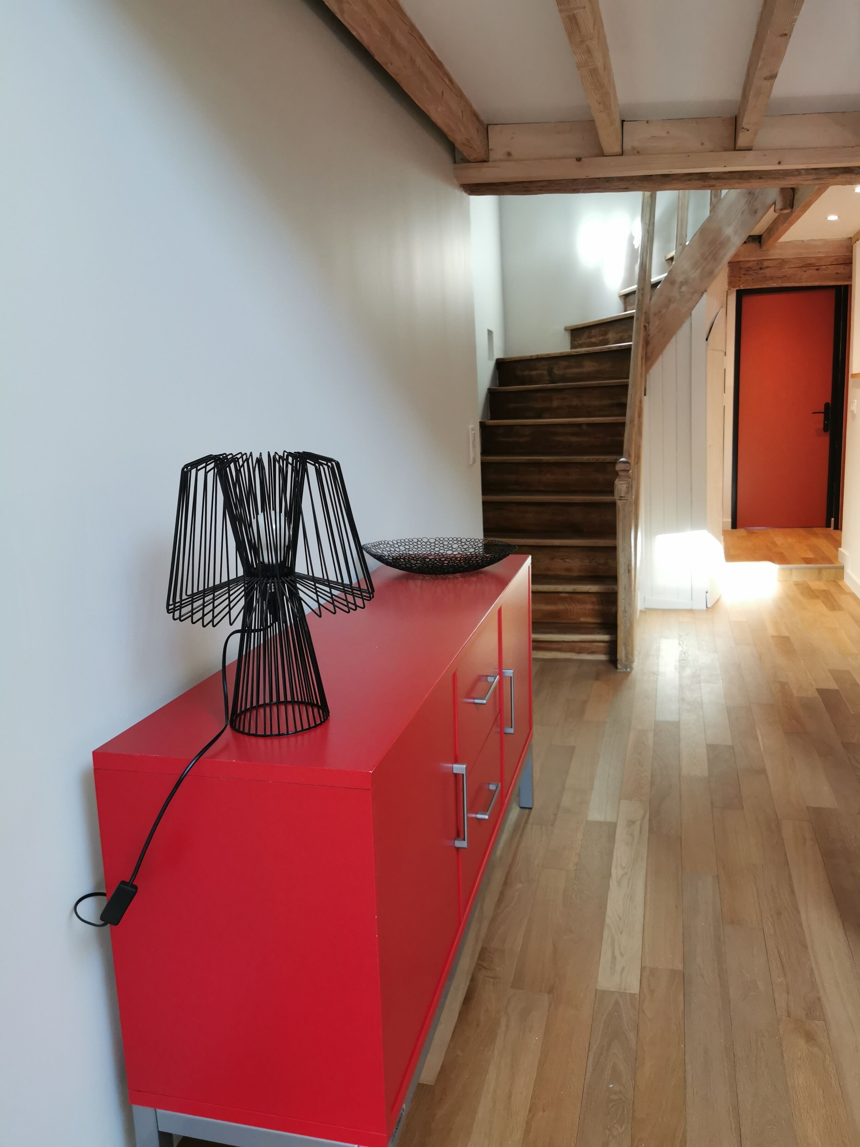 Een rode kast met lades, een zwarte lamp erop, een houten trap op de achtergrond en een rode deur aan het einde.