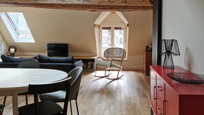 A spacious loft with a high ceiling, exposed wooden beams, a dining area with a round table and chairs, a rocking chair, and a red cabinet. - Old Annecy - 4-star apartment - near the lake - Pâquier - Historic center (Annecy)