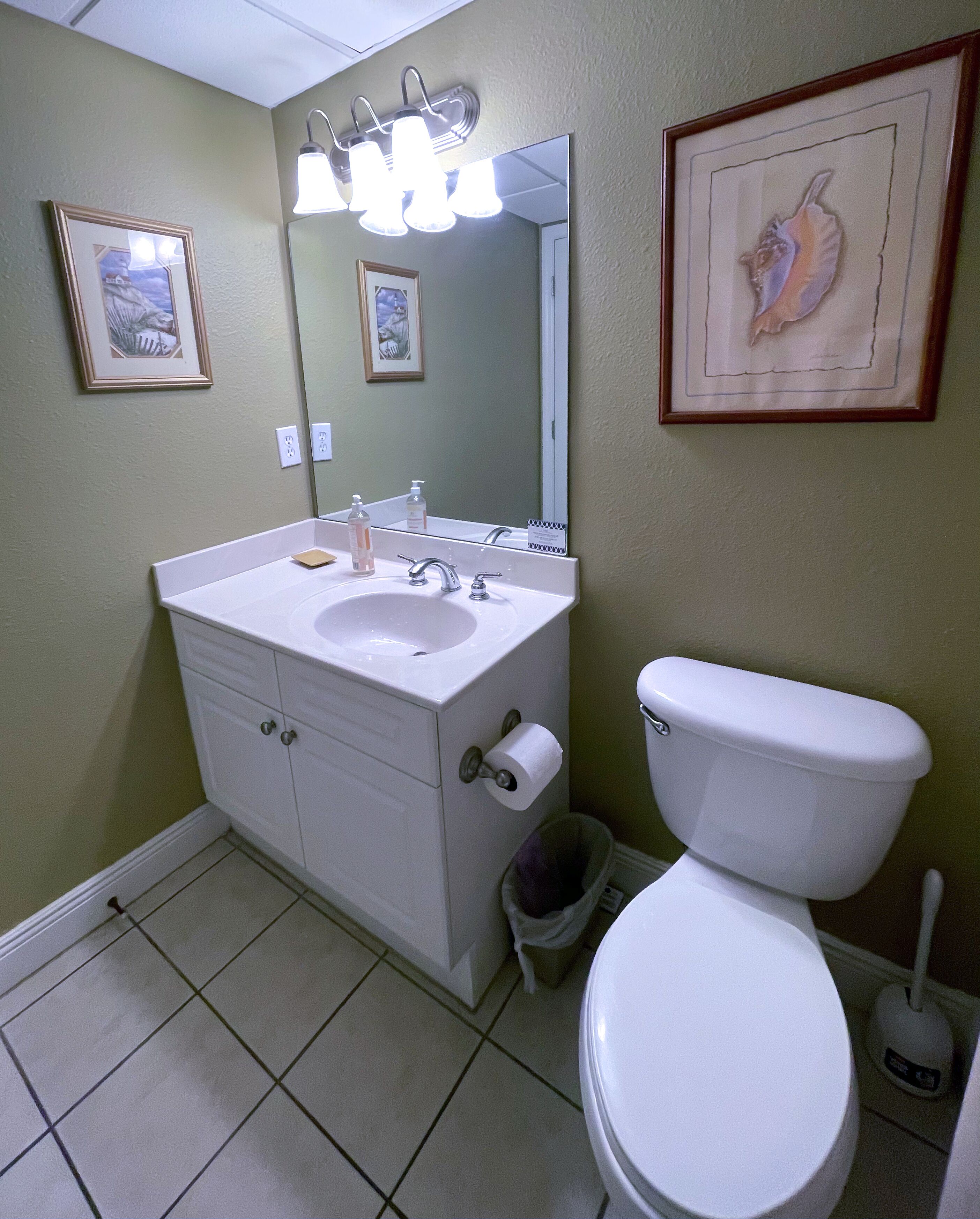 A bathroom with a white sink, mirror, and toilet. There are framed pictures on the wall and a trash can next to the toilet.