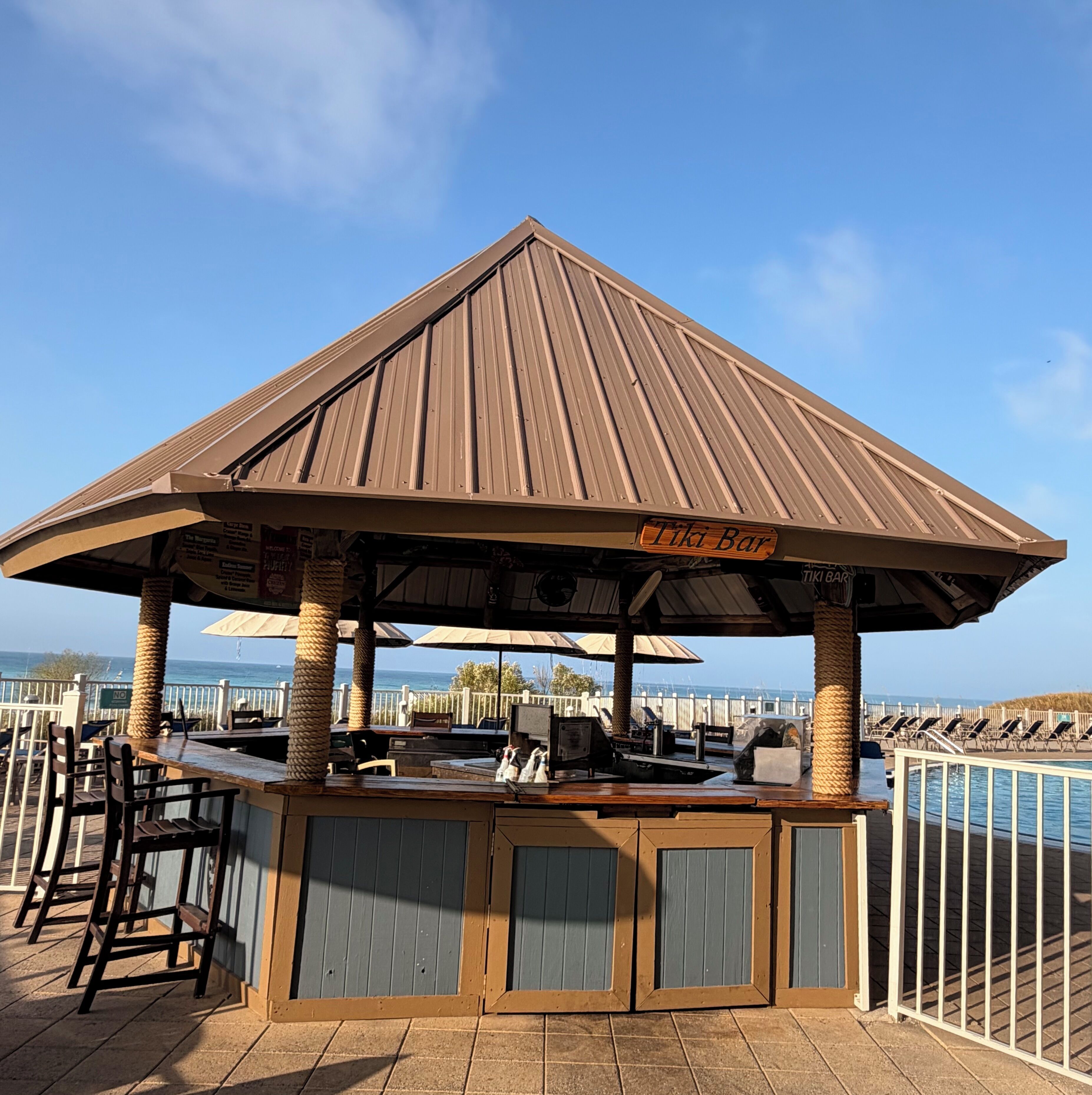 A beachside bar with a wooden roof, outdoor seating, and a view of the ocean.