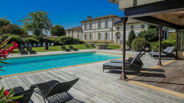 A wooden deck with a swimming pool, lounge chairs, and a large building in the background. - DOMAINE DE CEZAC-Proche Bordeaux-St-Emilion-Arcachon-Blaye (Cezac)