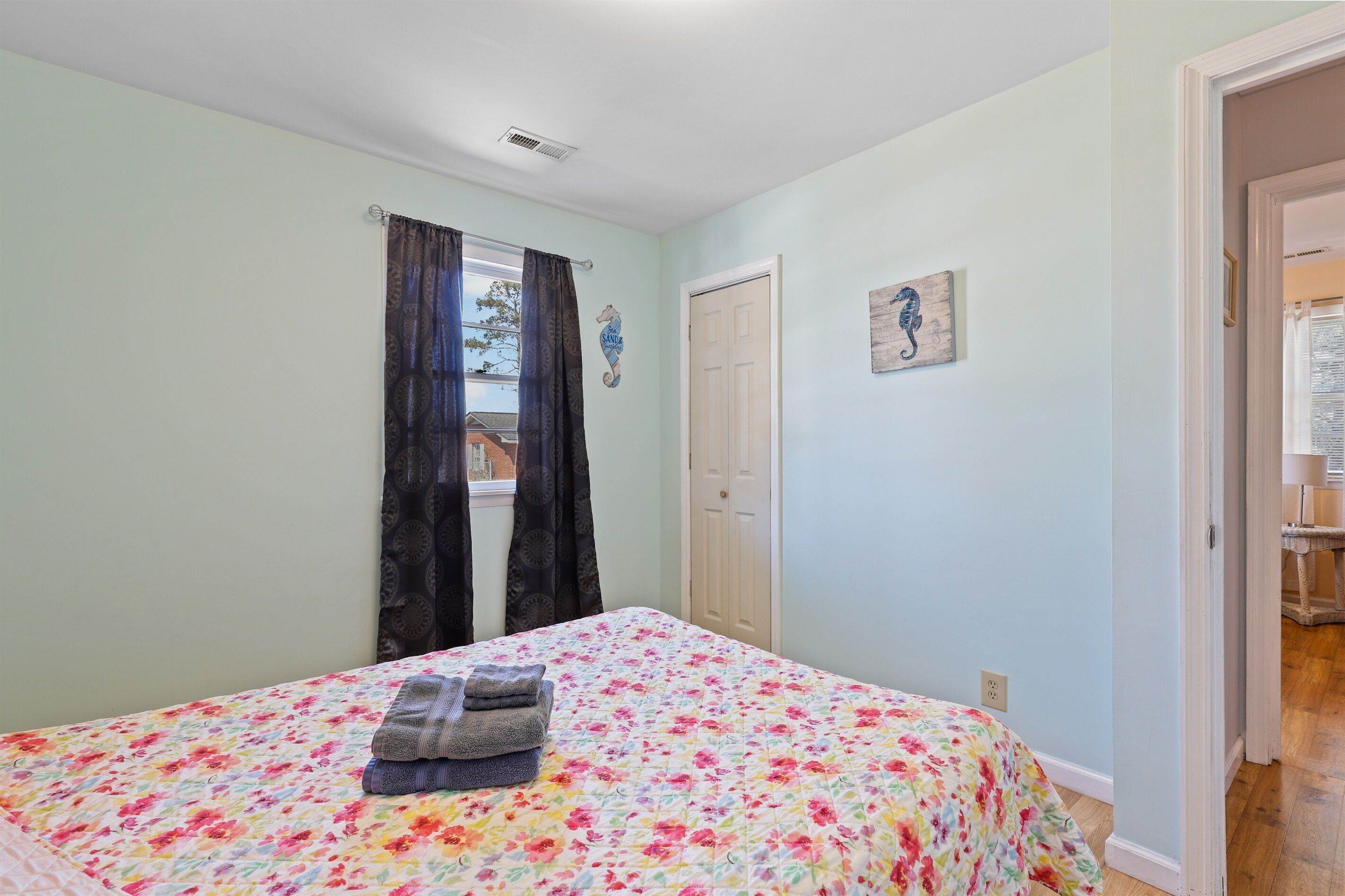 A bedroom with a floral bedspread, a window with curtains, a door, and a small artwork on the wall.