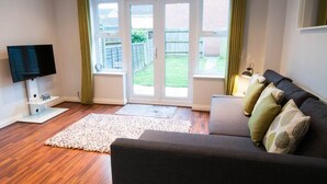 En stue med en mørk sofa, et fladskærms TV, en hvid måtte og udsigt til en baghave gennem en glasdør.