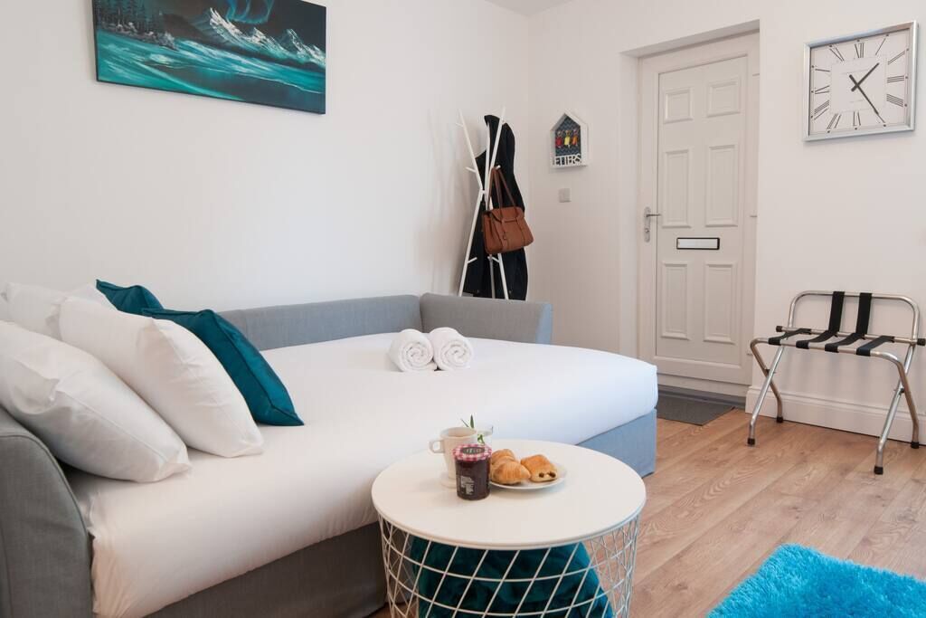 Une chambre d’hôtel moderne avec un lit, une petite table ronde sur laquelle sont posés des croissants, une horloge et un porte-manteau.