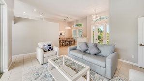 Ein modernes Wohnzimmer mit einem grauen Sofa, weißen Sesseln und einem Couchtisch. Im Hintergrund ist ein Essbereich mit Tisch und Stühlen zu sehen.