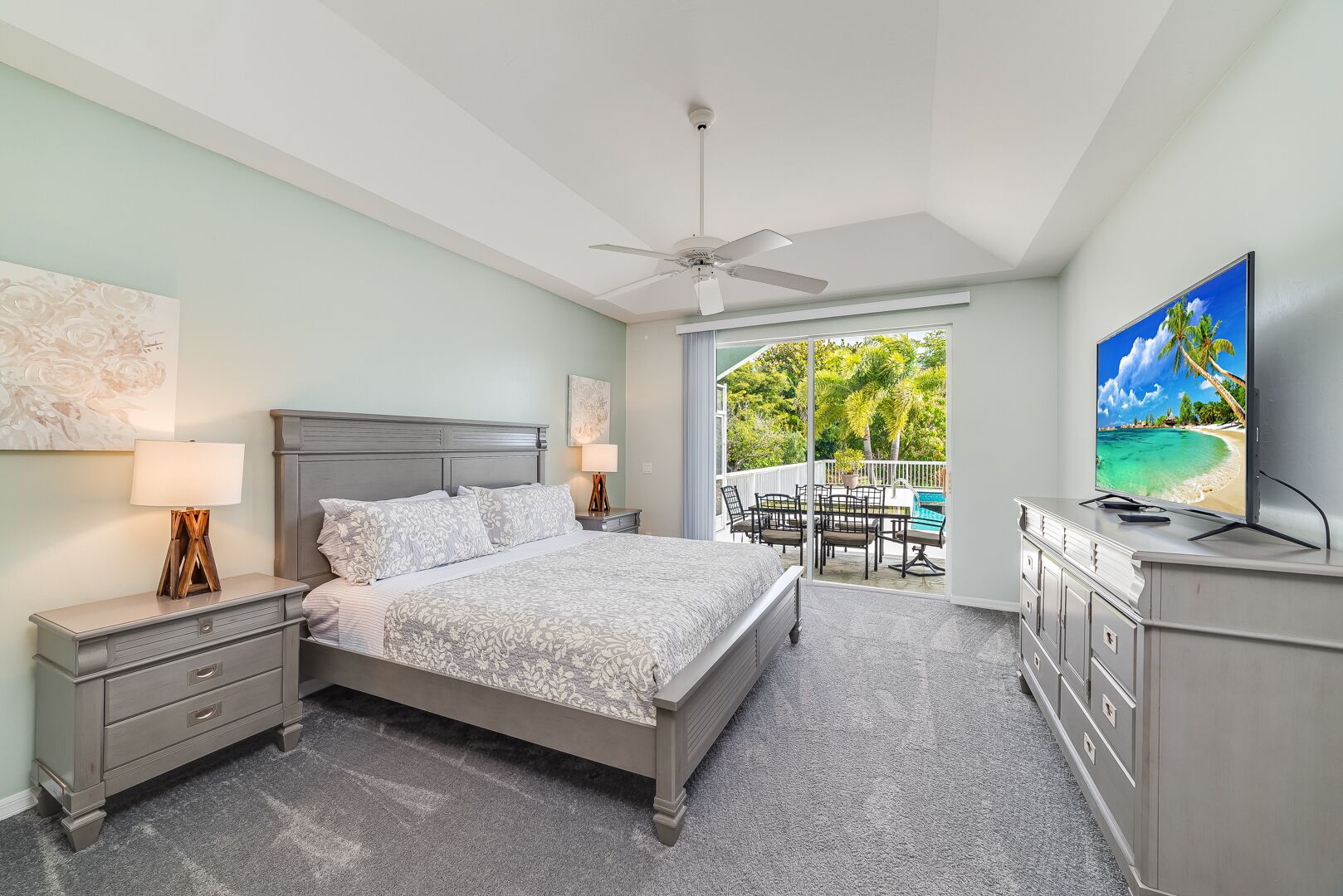 Ein Schlafzimmer mit Bett, Nachttischen, einem Schminktisch, einem Deckenventilator und einem Balkon mit Blick auf einen Strand.