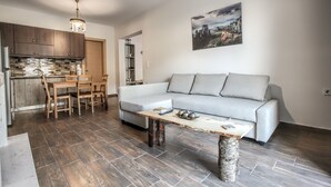 A modern living space with a grey sofa, wooden dining table, and chairs. A coffee table with decorative items is in the center. The kitchen has wooden cabinets and a tiled backsplash. - Terra di Meteora Suites 2 (Kalabaka)