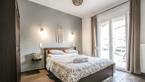 A bedroom with a large bed, a macrame wall hanging, and a view of the street through the window. - Terra di Meteora Suites 2 (Kalabaka)