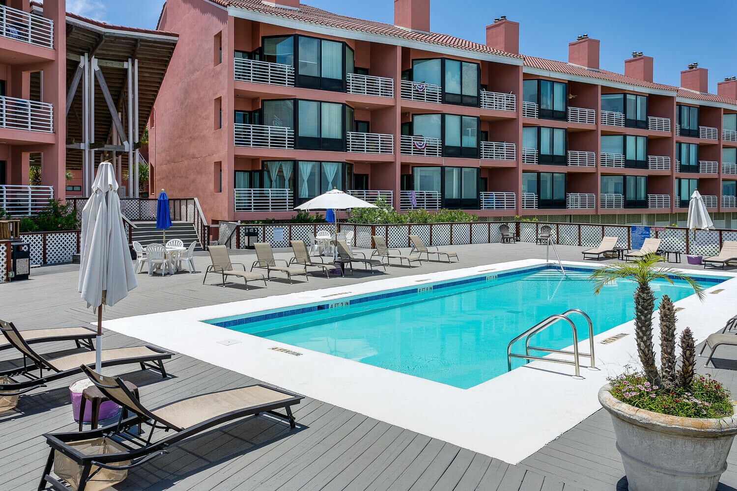 A pool area with lounge chairs, umbrellas, and a building with balconies in the background.