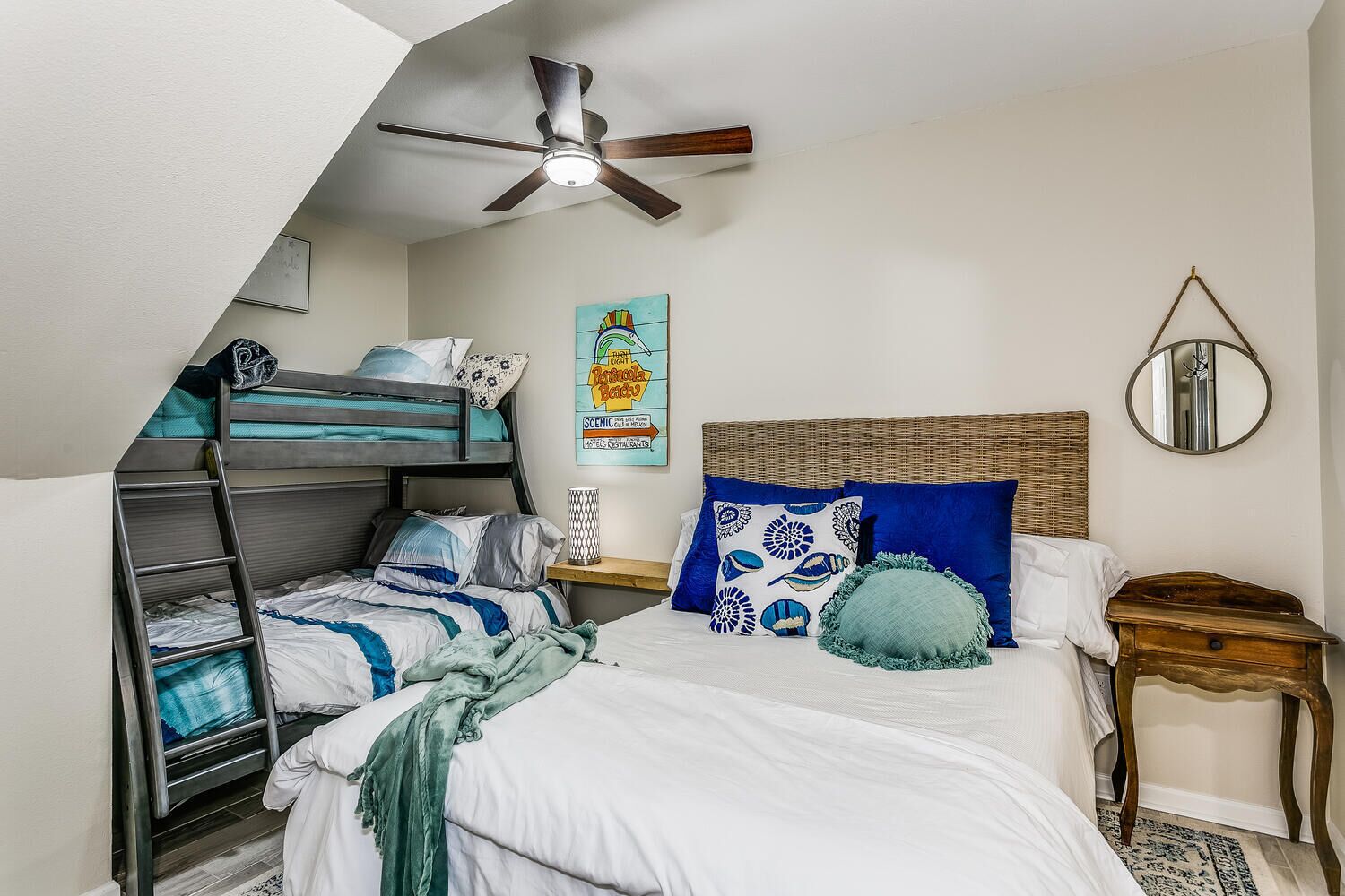 Une chambre de type dortoir avec des lits superposés, un ventilateur de plafond, un lit avec une literie bleue et blanche, une petite table de chevet avec une lampe et un miroir fixé au mur.