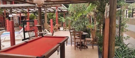 Una mesa de billar con superficie de fieltro rojo, rodeada de sillas de madera y una mesita.
