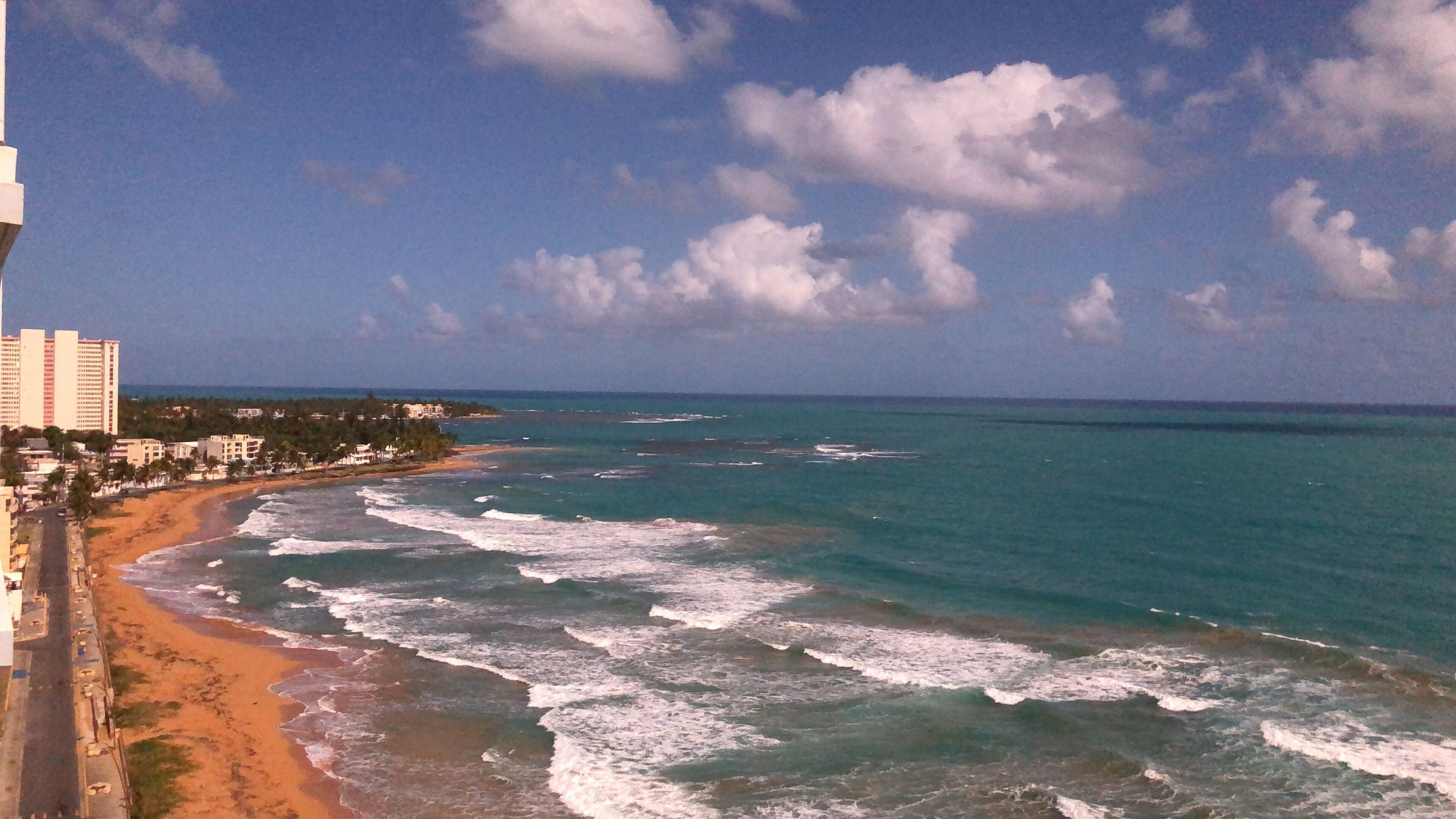 A beach with waves, a clear sky, and buildings in the background.