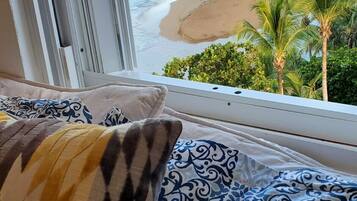 A bed with patterned pillows and a view of a beach with palm trees.