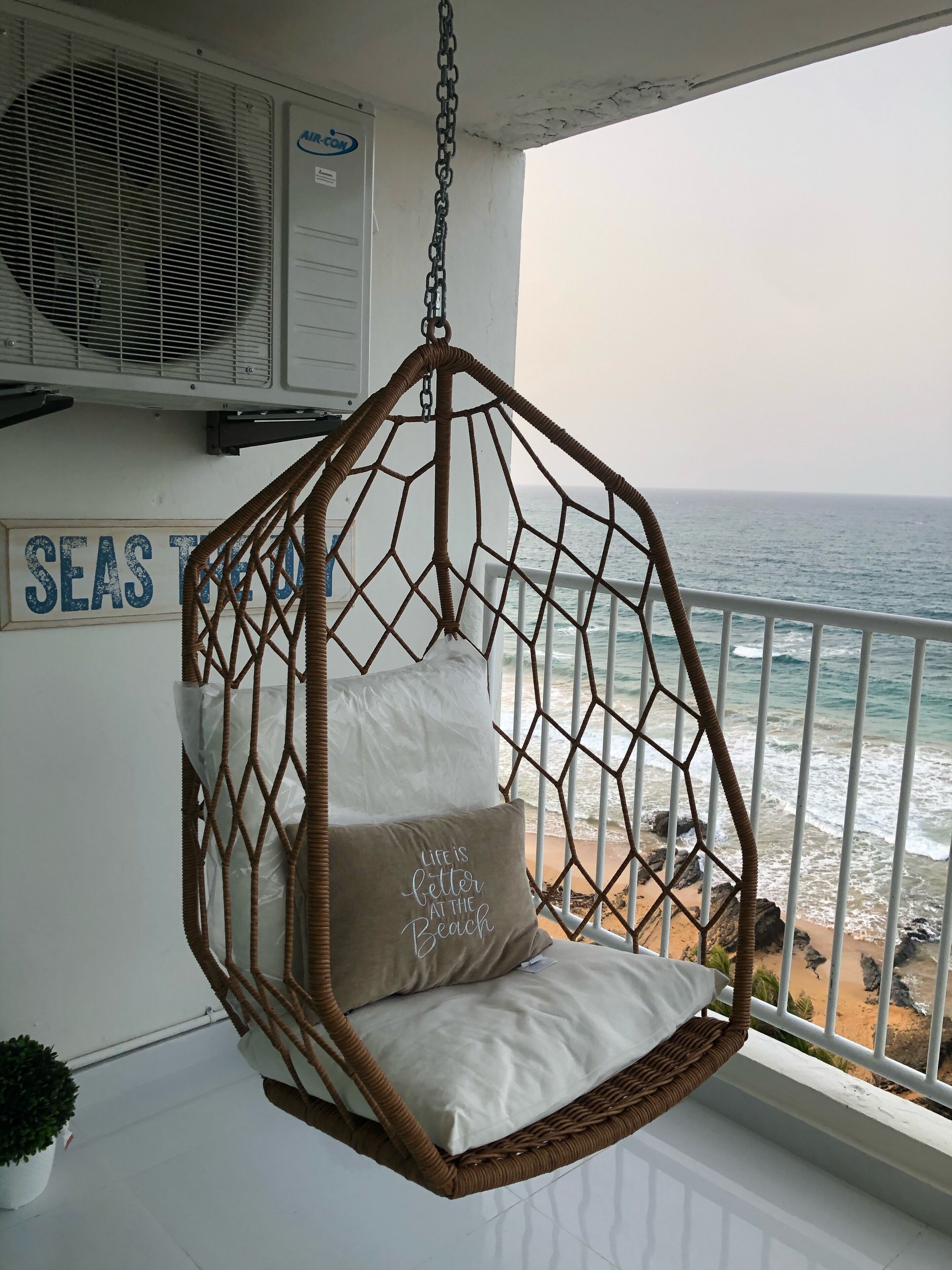 Una silla colgante de mimbre con cojines, un letrero que dice "SEAS TIDE" y vista al océano.