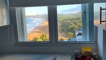 A kitchen with a large window overlooking a beach and greenery.