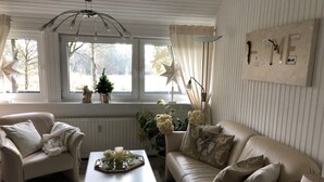 Un soggiorno accogliente con un divano bianco, una poltrona beige e un tavolino da caffè bianco. Si nota una finestra con tende, un termosifone e una targa appesa al muro con la scritta “HOME”.
