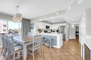 A modern kitchen with white cabinets, a central island, and a dining area with a wooden table and chairs. - Oceanfront Luxury Top Location Lazy River Pool Grills (North Myrtle Beach)