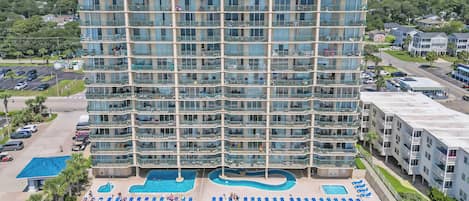 A high-rise hotel with multiple balconies, a swimming pool, and a well-maintained lawn.