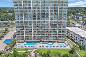 A high-rise hotel with multiple balconies, a swimming pool, and a well-maintained lawn. - Oceanfront Luxury Top Location Lazy River Pool Grills (North Myrtle Beach)