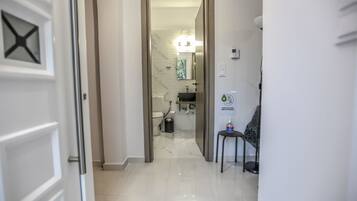 A hallway leading to a bathroom with a toilet, sink, and a trash bin. There is a chair and a bottle of hand sanitizer on a small table.