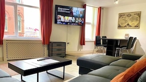 Ein modernes Wohnzimmer mit einer schwarzen Couch, einem Couchtisch, einem an der Wand befestigten Fernseher, einem kleinen Schreibtisch mit Stühlen und einer Heizung.