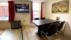 Ein Zimmer mit einem großen Flachbildfernseher an der Wand, der eine Netflix-Oberfläche zeigt, einem Holztisch mit vier Stühlen, einem Fenster mit roten Vorhängen und einem Wandbild einer Weltkarte.