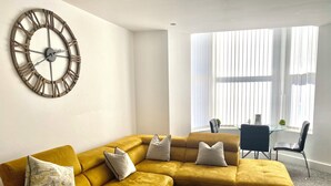 Una sala de estar con un sofá modular color mostaza, un reloj de pared grande y un comedor con una mesa de vidrio y sillas.