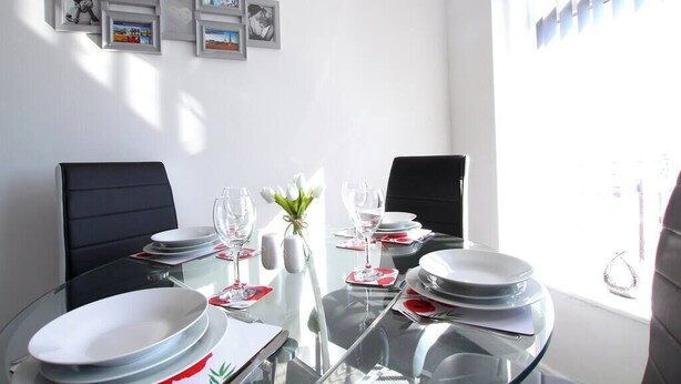 A dining area with a glass table set for two, two black chairs, a vase with white flowers, and framed pictures on the wall.