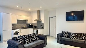 A modern living room with two sofas, a glass coffee table, and a kitchen area with a washing machine.