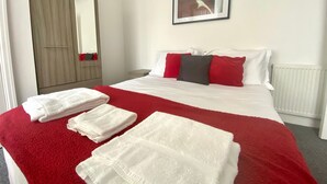 Habitación de hotel con cama, ropa de cama blanca, almohadas rojas y negras, una manta roja y un radiador blanco.