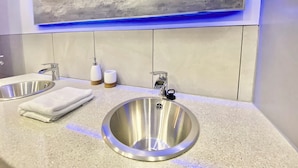A modern bathroom with a large, round stainless steel sink, a mirror, and a wall-mounted soap dispenser.