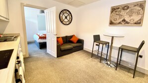 A compact living space with a kitchenette, a sofa with orange cushions, a round wall clock, a small table with two chairs, and a world map on the wall. - Winter Gardens Service Apartment 2 (Blackpool)