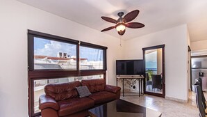 A living room with a leather sofa, a TV, a ceiling fan, and a view through a large window. - Nice Caribbean style apartment in beachfront condominium (Playa del Carmen)