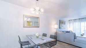 Una sala de estar moderna con una mesa de comedor de vidrio, sillas transparentes, un sofá gris, una araña y un ventanal con cortinas.