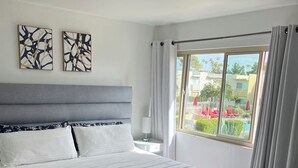 Habitación de hotel moderna con una cama grande, un cabecero gris y vistas a una piscina y edificios.