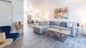 Una sala de estar moderna con un sofá seccional gris, un sillón azul y una mesa de centro. Hay una alfombra estampada en el suelo, una lámpara sobre una mesita auxiliar y un cuadro en la pared.