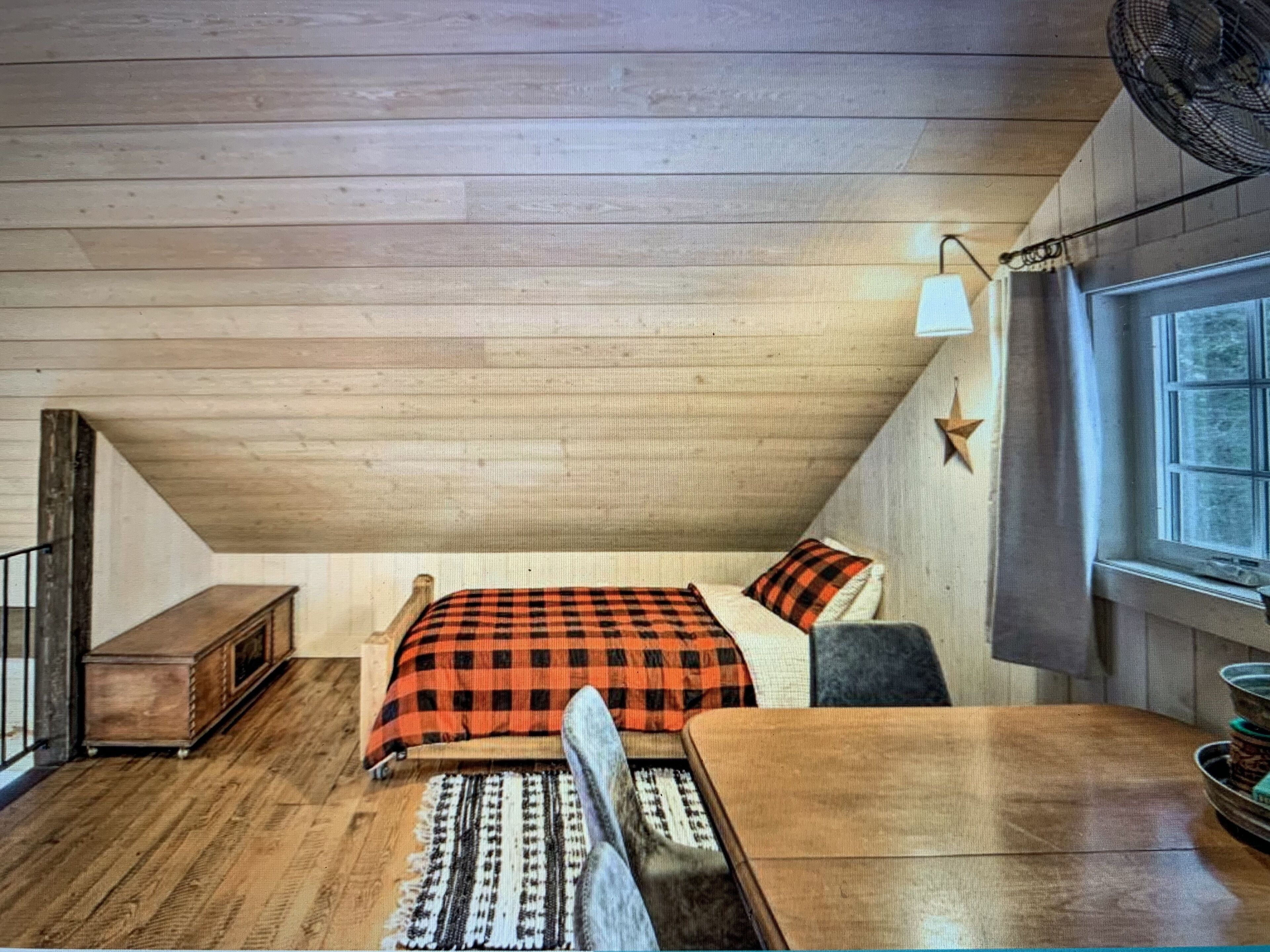 Ein gemütliches Schlafzimmer im Dachgeschoss mit Holzbalkendecke, einem Bett mit kariertem Überwurf, einem kleinen Nachttisch aus Holz und einem Fenster mit Vorhängen.