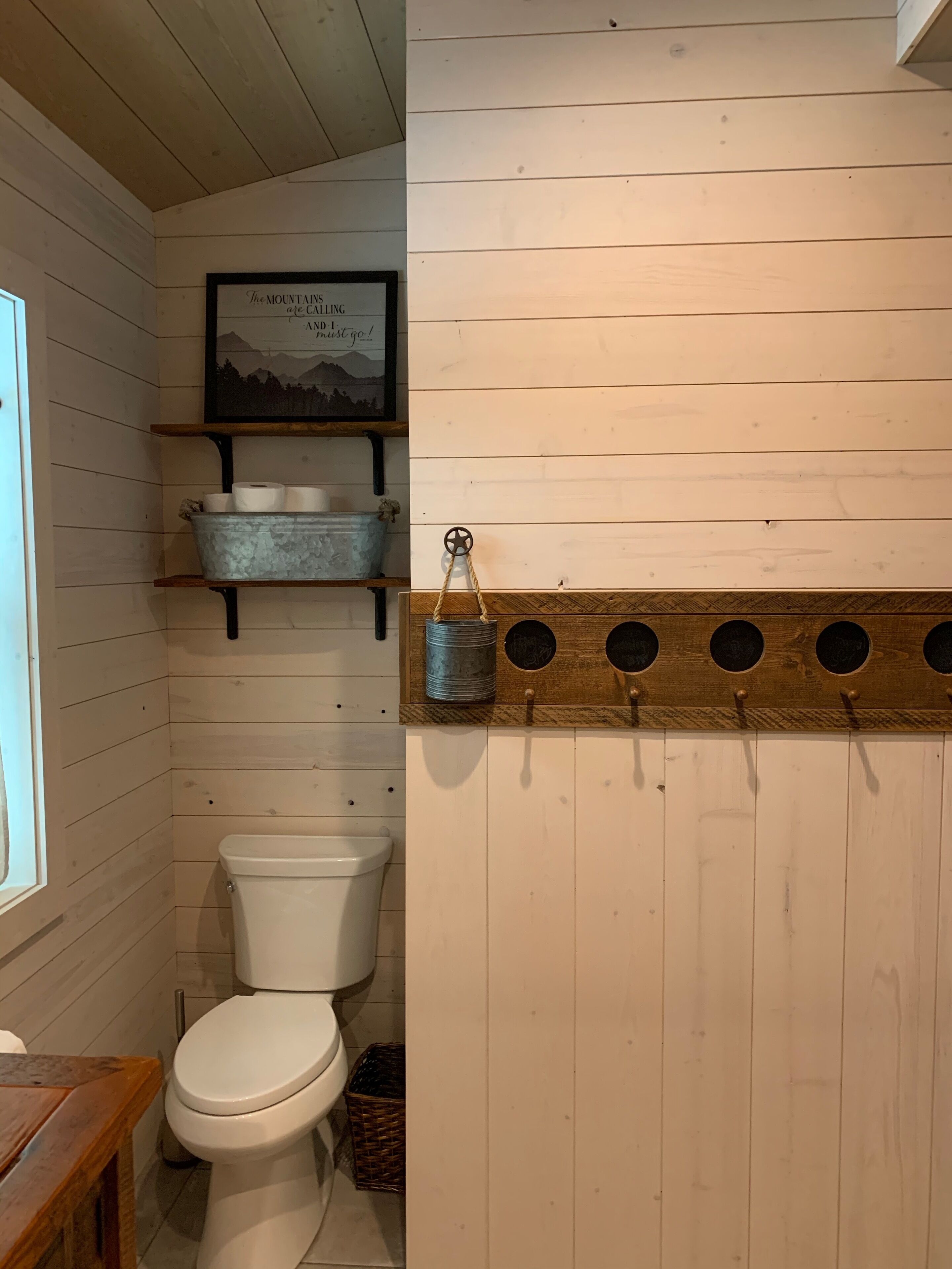 A small, rustic bathroom with wooden walls, a white toilet, a shelf with metal containers, and a framed quote on the wall.