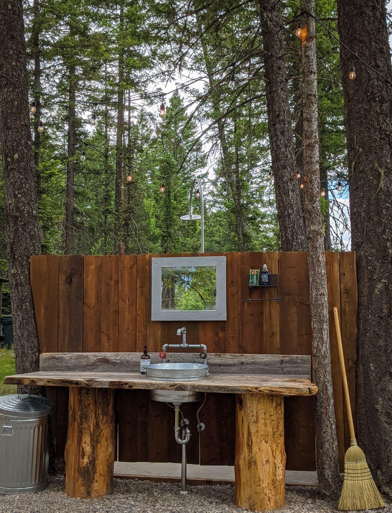 An outdoor wooden bench with a metal sink, surrounded by trees and string lights.